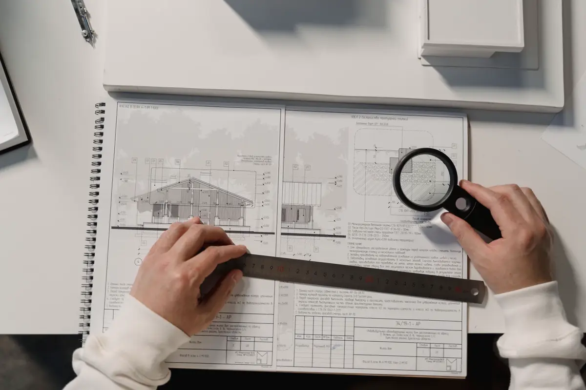 Mãos com régua e lupa analisando a planta baixa e a fachada de uma edificação