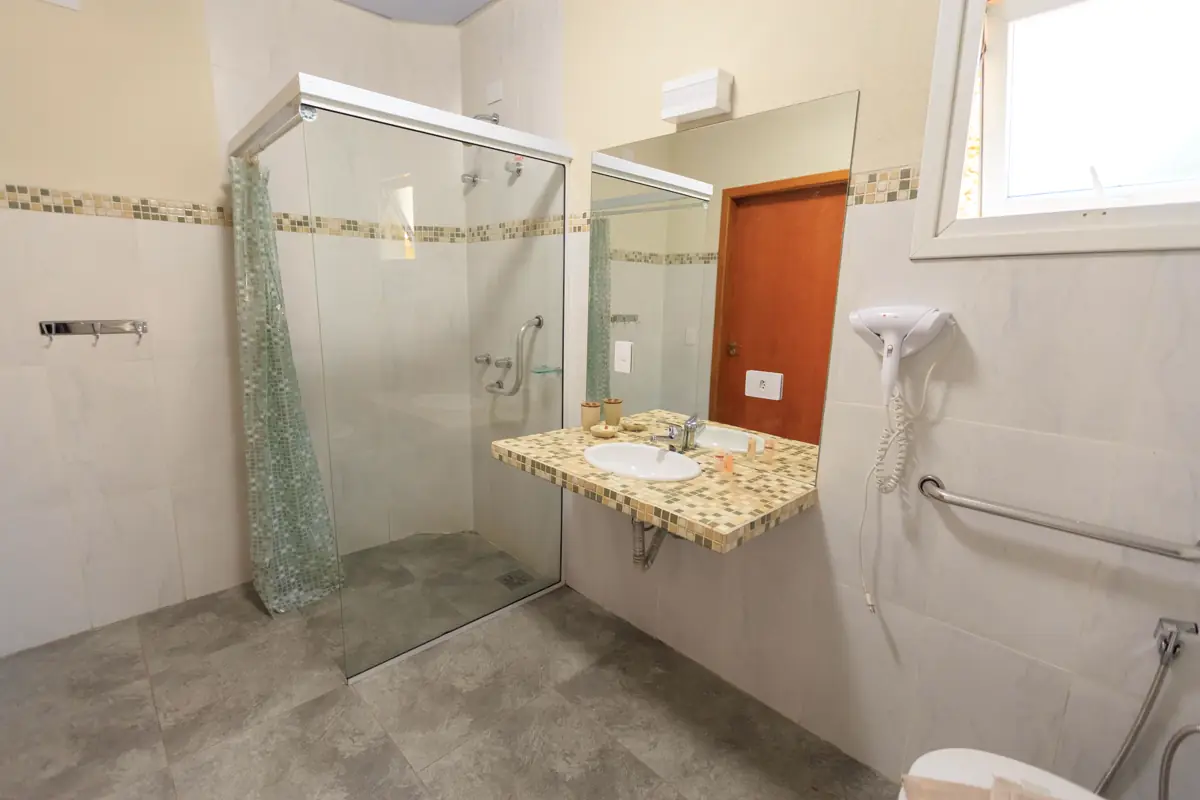 Banheiro com box de vidro, barra de apoio na parede, bancada de granito e piso cinza