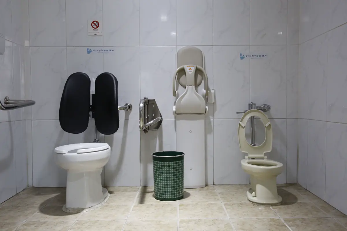 Sanitário público com bacias, barras de apoio rebatíveis e acessórios nas paredes revestidas