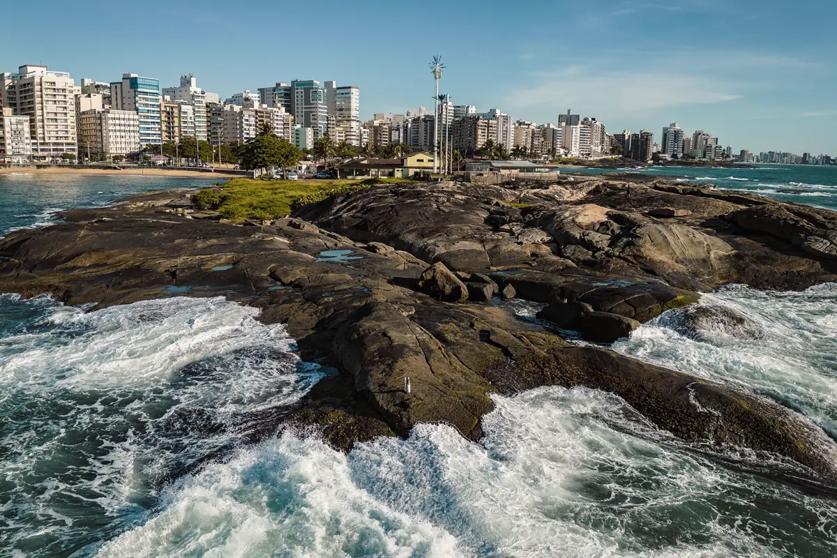 Costão rochoso com ondas quebrando e a fileira de prédios da orla de Guarapari ao fundo