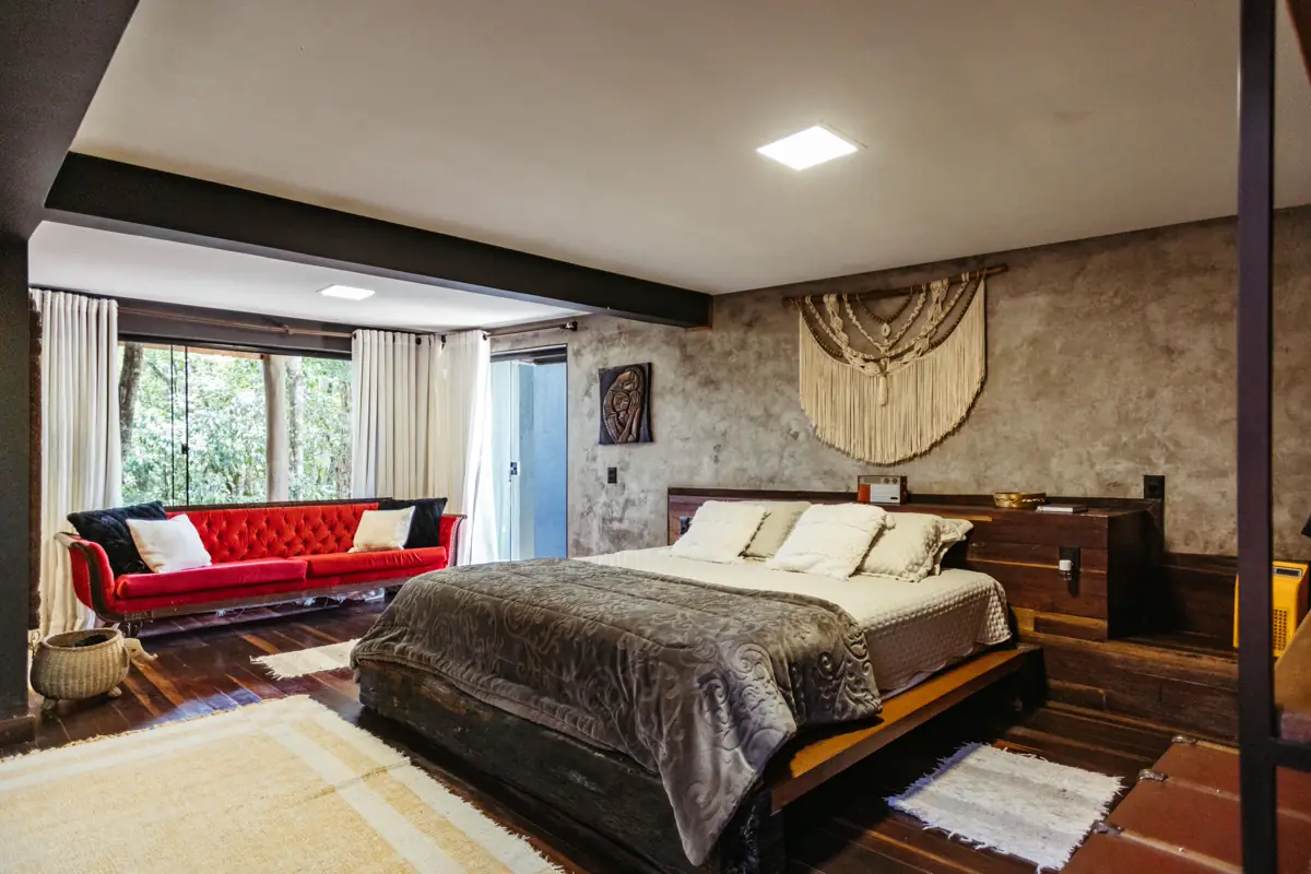Quarto de pousada amplo com cama de casal, sofá vermelho e janelas para a mata