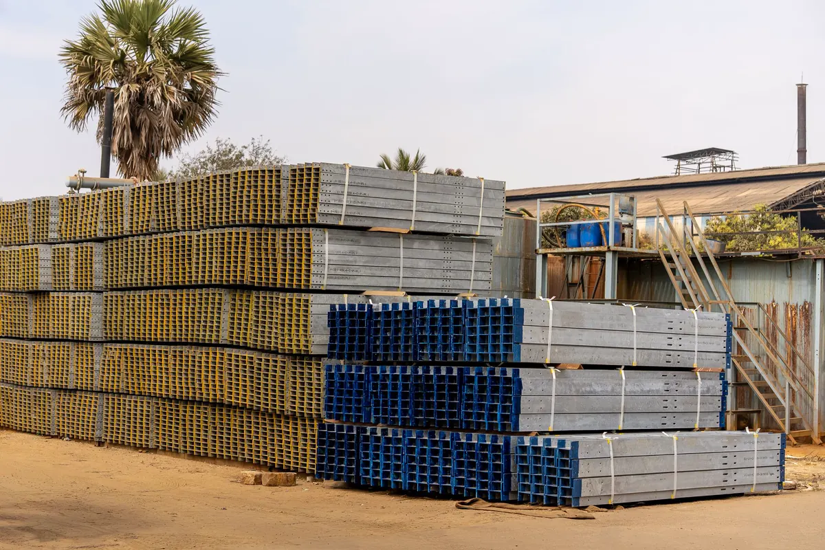 Feixes de perfis de aço galvanizados e perfurados, cintados e empilhados em pátio aberto, com as pontas pintadas de amarelo e de azul, ao lado de um galpão e de uma palmeira
