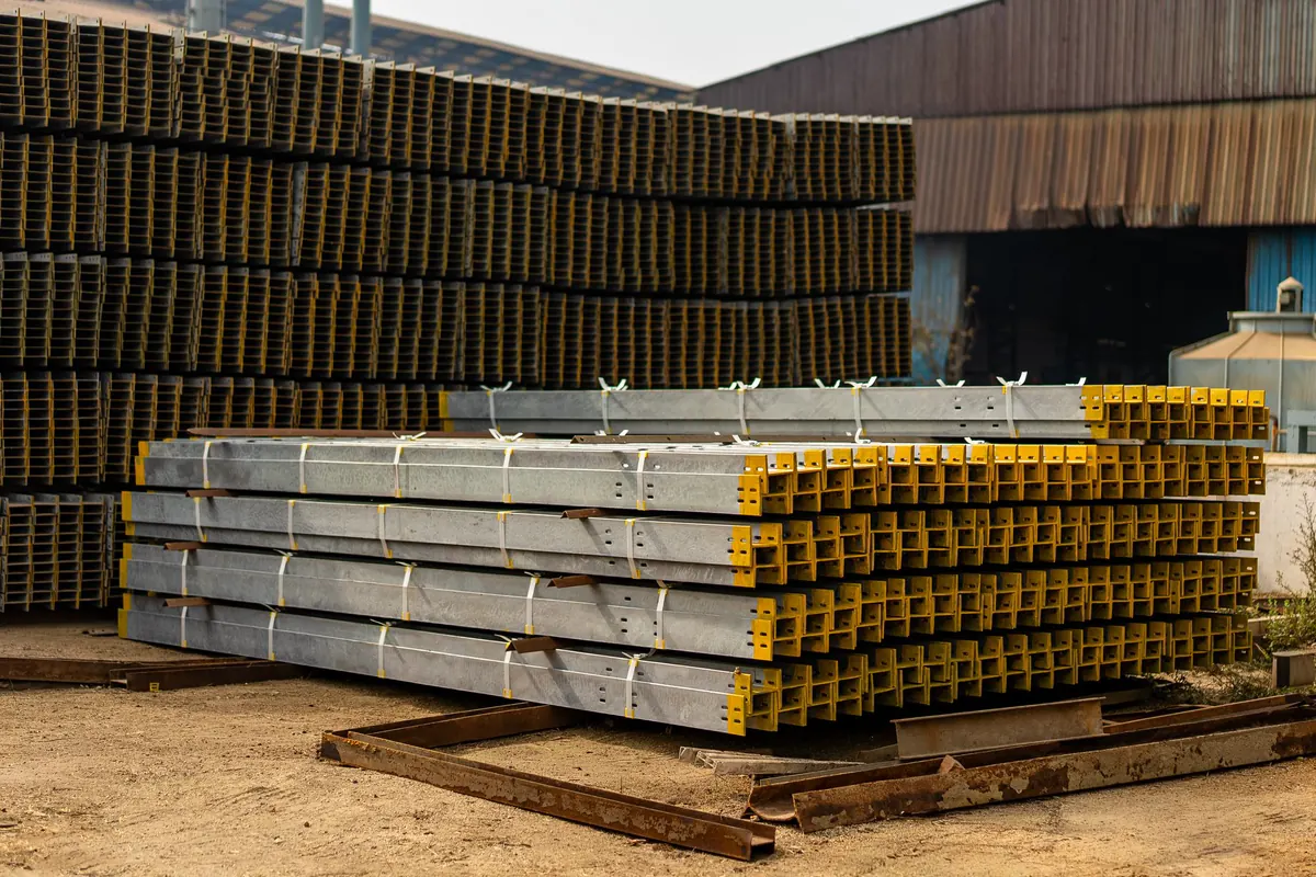 Feixes de perfis de aço galvanizados com as pontas pintadas de amarelo, cintados e apoiados sobre calços, diante de grandes pilhas de perfis escuros em pátio de chão batido