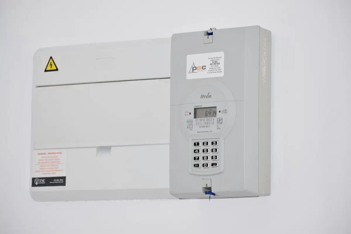 Medidor de energia eletrônico ao lado de uma caixa de disjuntores numa parede branca