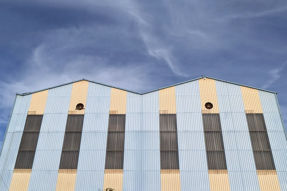 Fachada de dois galpões geminados de duas águas, com telha ondulada em faixas azul-claras e amarelas, painéis escuros de ventilação e dois exaustores circulares