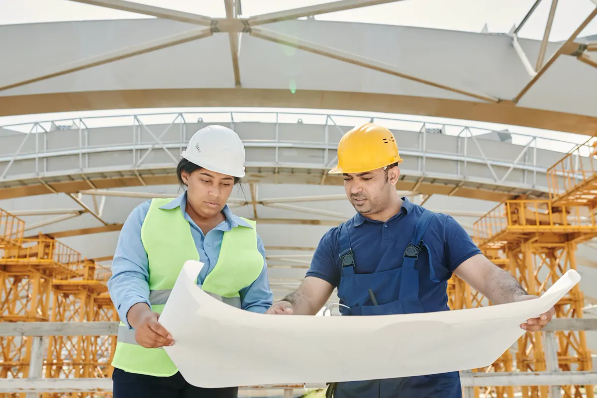 Engenheira de capacete branco e colete refletivo e engenheiro de capacete amarelo conferindo uma prancha de projeto sob estrutura de cobertura em treliça metálica em construção