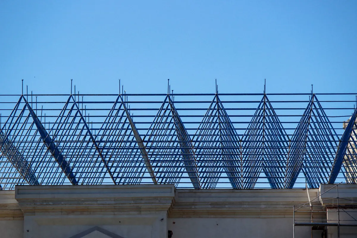 Cobertura em treliças metálicas azuis montada sobre o topo de um edifício de alvenaria clara, com andaime na lateral e céu azul ao fundo
