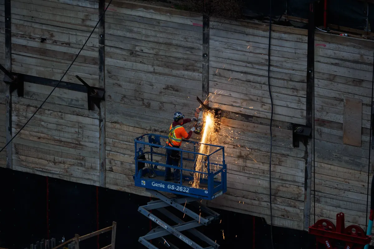 Operário de capacete e colete refletivo sobre plataforma elevatória tipo tesoura, cortando peça metálica com faíscas