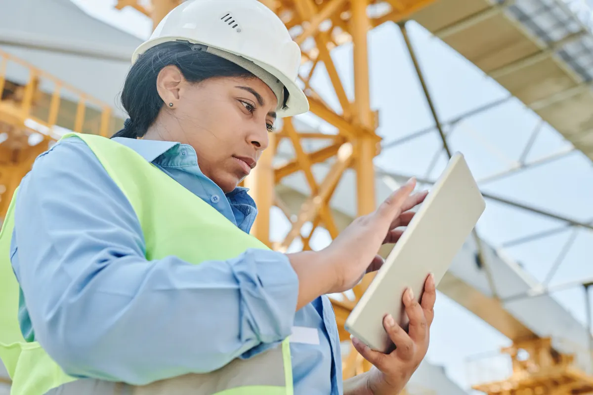 Engenheira de capacete branco e colete refletivo consultando um tablet diante de estrutura metálica amarela em construção