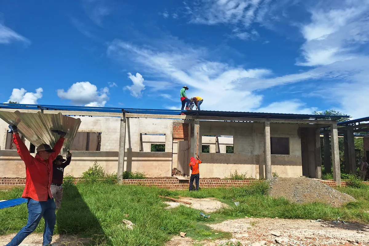 Equipe de obra carregando telhas metálicas até uma edificação em construção, com dois trabalhadores fixando a cobertura sobre as terças