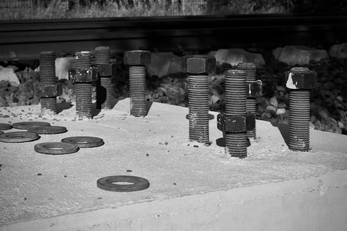 Chumbadores roscados com porcas saindo de um bloco de concreto, com arruelas soltas sobre a superfície, em foto preto e branco