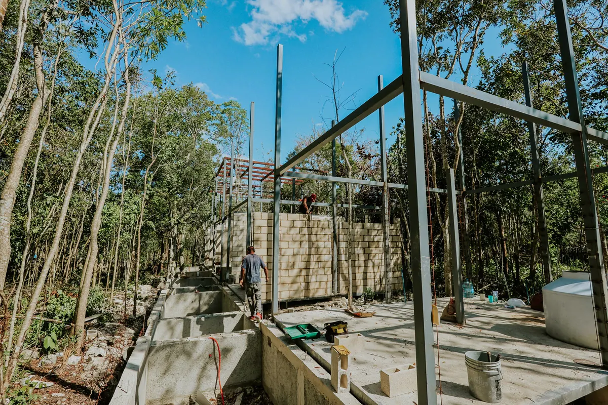 Pilares e vigas metálicas cinza montados sobre base de concreto em obra cercada de árvores, com um trabalhador caminhando sobre a viga de fundação e outro no alto da parede de blocos