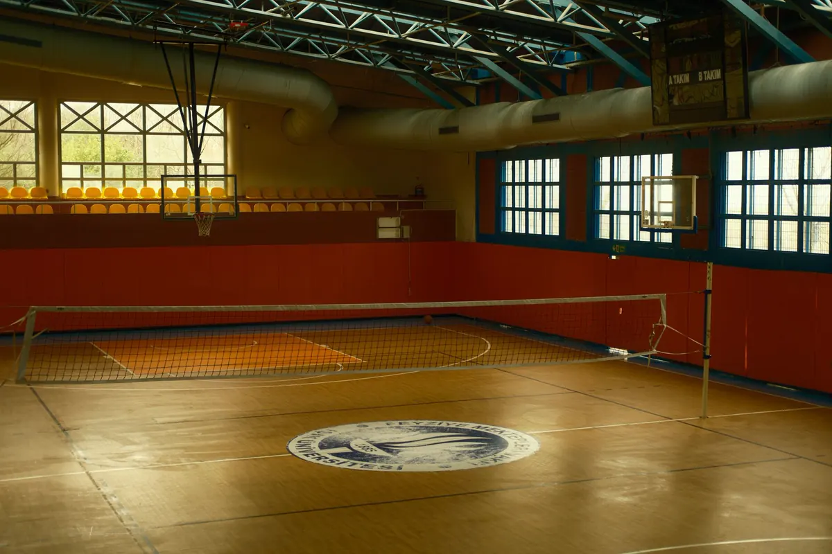 Quadra coberta de ginásio com piso de madeira, rede de vôlei armada, tabelas de basquete e treliças metálicas aparentes sob a cobertura