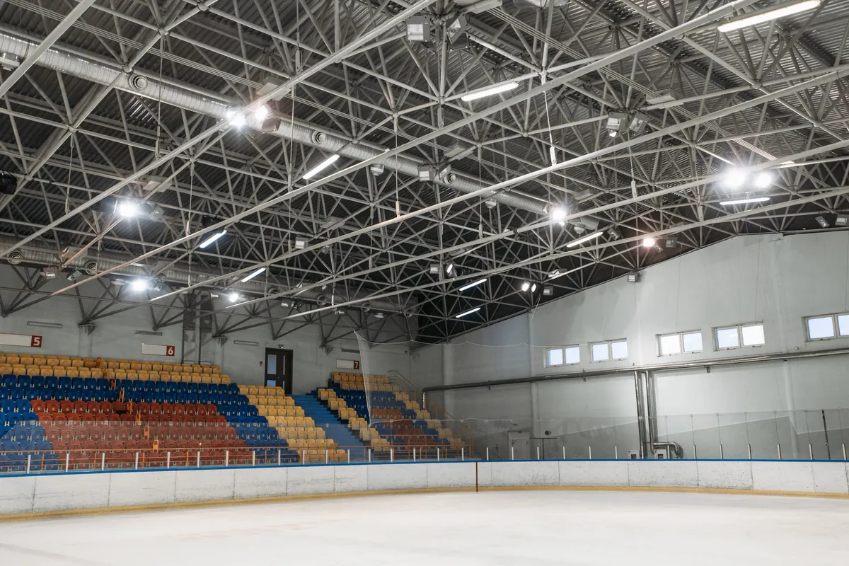 Interior de ginásio com cobertura em treliça espacial metálica de grande vão, refletores presos à estrutura, arquibancada colorida e janelas altas na parede lateral