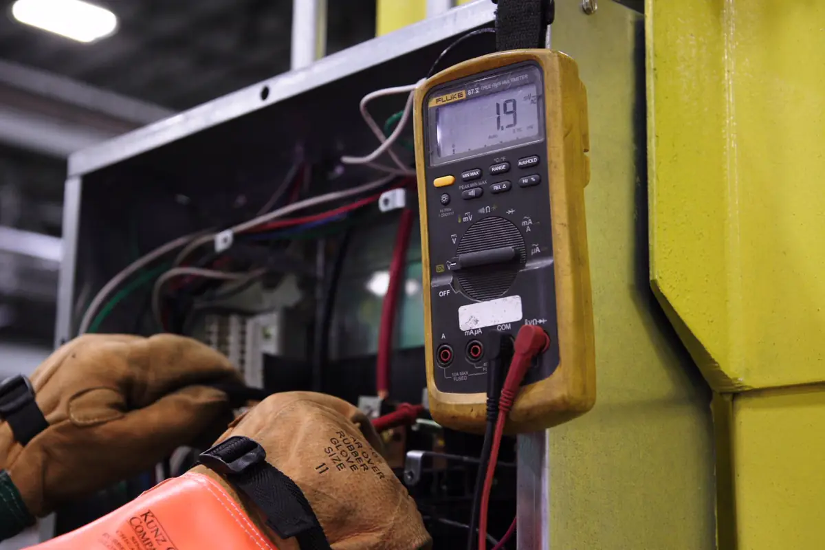 Multímetro digital com pontas de prova ligado aos cabos de um painel elétrico, manuseado com luvas de couro