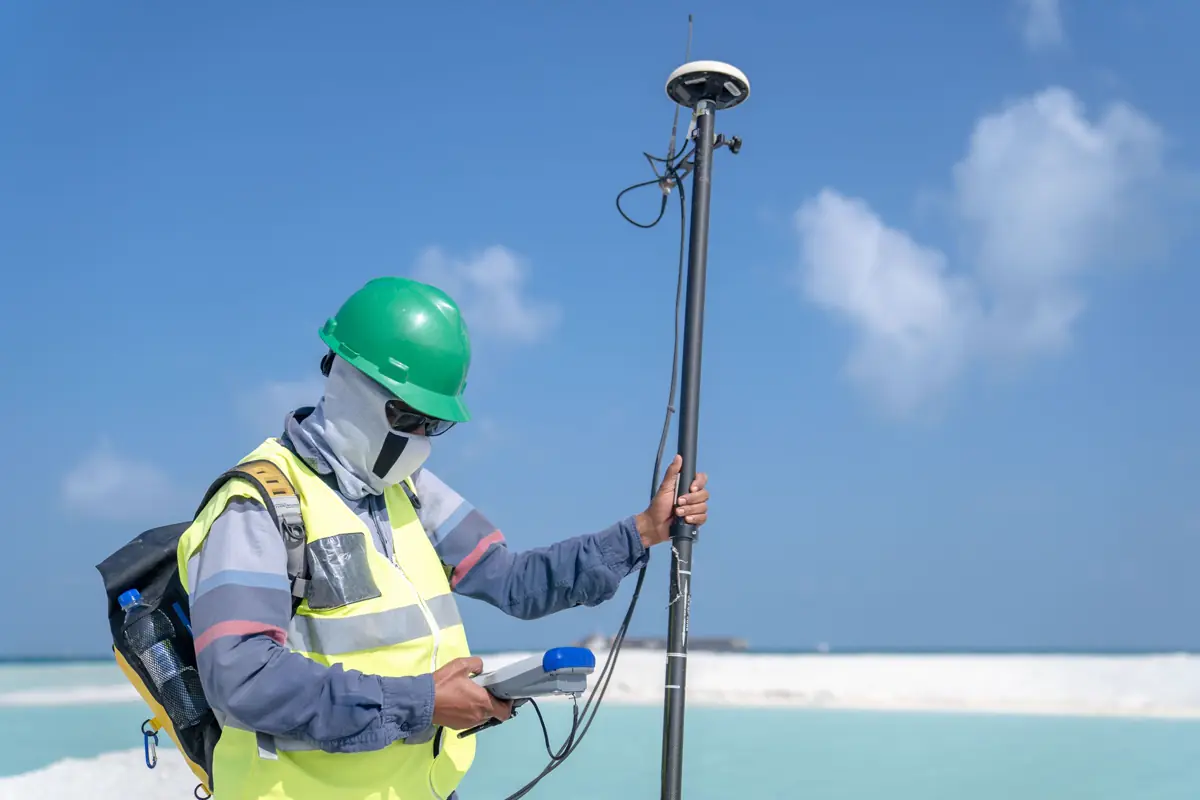 Técnico de capacete e colete segurando bastão com receptor GNSS e coletora de dados numa área de praia